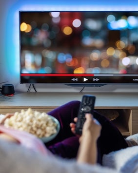 A person holding a remote control with a focus on a bowl of popcorn, with a blurred television screen in the background.