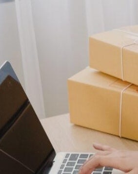 The image shows a close-up of a person's hand typing on a laptop keyboard, with a stack of brown cardboard boxes next to it.